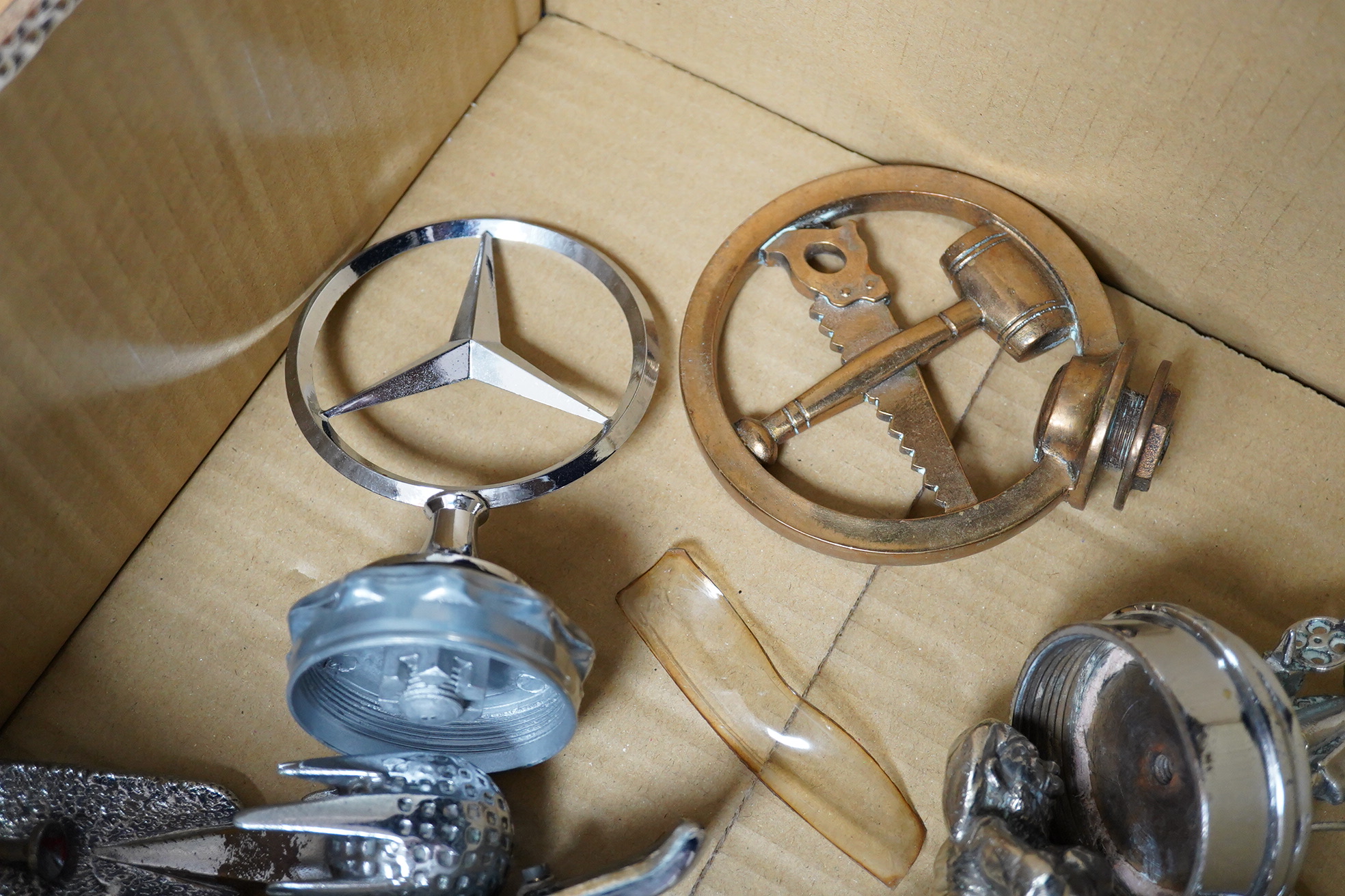 Seven chrome and brass car mascots, including a Mercedes-Benz logo, a figure of a blacksmith, a caricature figure on a beer barrel, a golf ball in mid flight, etc. together with a chromed housing unit for an electric lam